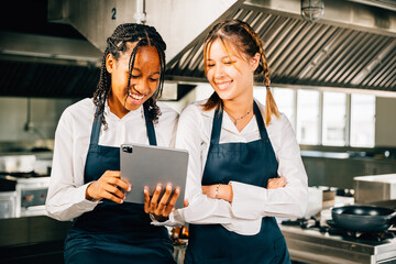 In a professional restaurant kitchen two famous chefs discuss video blog using a tablet. Modern technology aiding discussion colleagues smiling searching reading and connecting.