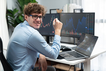 Profile of smiling young stock trader with curly hair looking at camera, sitting against on financial exchange screen at modern office. Concept of dynamic business digital investment. Gusher. - obrazy, fototapety, plakaty