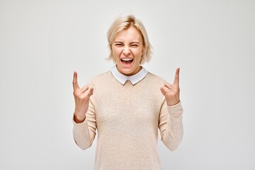 Excited woman with blonde hair making rock and roll hand gesture, wearing a casual sweater,...