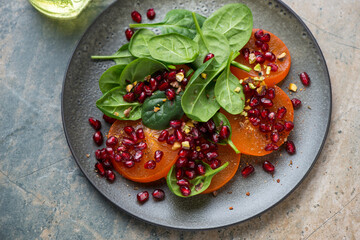 Plate with persimmon, fresh spinach and pomegranate salad, horizontal shot on a beige and grey granite surface, elevated view