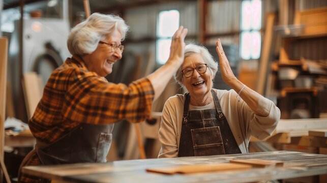 Senior craftswomen giving a high-five in a workshop. Photo depicts joyous moment between two elderly women wearing casual clothing and aprons, surrounded by woodwork tools.