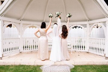 Beautiful bride with bridesmaids standing outdoors in a green garden, women raising hands with white flower bouquets in celebration of wedding day, happy bridal party 
