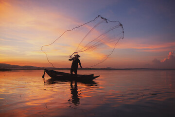 Fisherman casting his net on during sunset. Silhouette Asian fisherman on wooden boat casting a net for freshwater fish. Thailand culture.