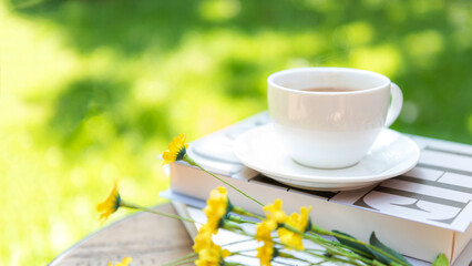 White steaming cup of hot coffee owith yellow flowers and green nature blurred background.