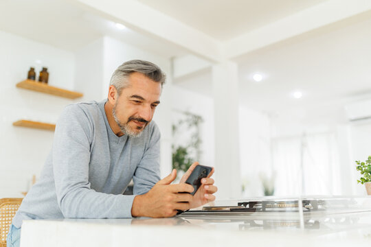 Confident mature man with a charming smile leaning on kitchen counter while texting on his smartphone in a bright home