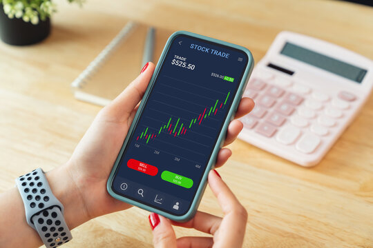Female trader using smartphone with stock market investments and graph on screen to analyze trading data, buy or sell and checking price on mobile application.