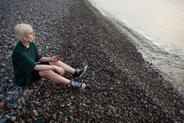 Melancholy young girl with short blonde bob hair and green dress on cold sea beach