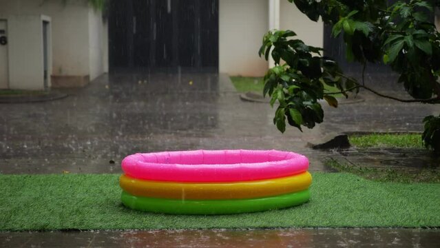 In front of suburban house, empty kiddie pool sits under pouring tropical rain. Surroundings are soaked, and warm rain provides refreshing relief on hot day. Empty street and house on background