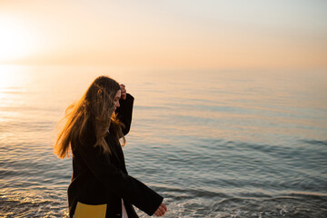 Stylish young woman wearing trendy jacket and dress walking at sea coast line over sunset outdoor. Spring season.