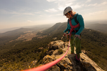 A woman is standing on a mountain with a red rope tied to her. She is wearing a helmet and a blue...