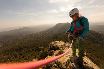 A woman is standing on a mountain top, wearing a harness and a helmet. She is preparing to rappel...