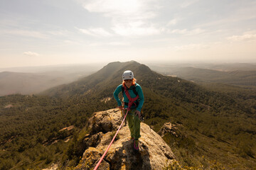 A woman is standing on a mountain top, wearing a helmet and harness. She is looking out over the...