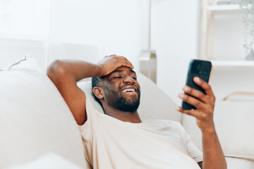 Happy African American man sitting on a black sofa, using his mobile phone He is casually dressed...