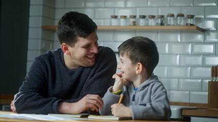Young father help schoolboy with homework at home. Dad helping to son with writing while sitting in the kitchen. Adult parent or tutor with kid boy learning.

