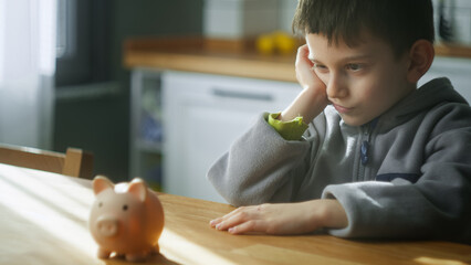 Bored cute boy sit in the kitchen thinking and looking to a piggy bank. Concept of teaching...