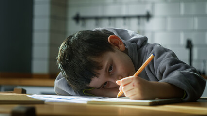Tired little schoolboy kid lying on table desk writing homework, boring class lesson drawing in...