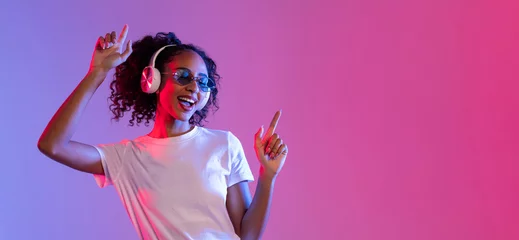 Zelfklevend Fotobehang Joyful black woman with sunglasses and headphones dancing on neon background, free space © Prostock-studio