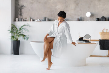 Black woman wearing bathrobe sitting on bathtub and touching her beautiful legs