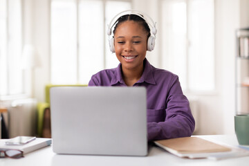Smiling female black student in headphones using laptop at desk at home
