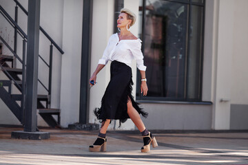 Confidence and sophistication mature woman walks city streets in fashionable ensemble chic white shirt and black fringed skirt with high heeled sandals and socks, fashion icon of style