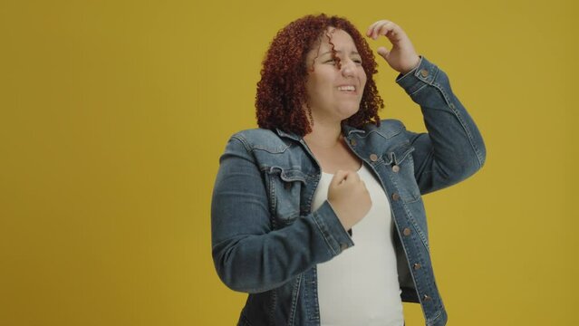 Smiling overjoyed hispanic young woman in denin jacket posing doing winner gesture, say Yes isolated on yellow background in studio. People sincere emotions, lifestyle concept
