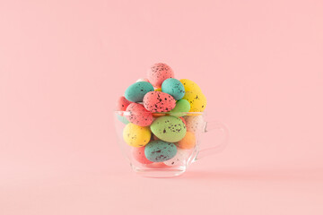 A glass cup full of multi-colored Easter egg on a pastel pink background with copy space. 
