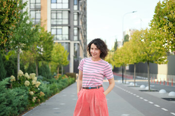 Happy young woman walking outside. One confident girl walks outdoors in street smiling. Urban city, residential complex, real estate concept. Brunette female adult in pink clothes. High quality photo