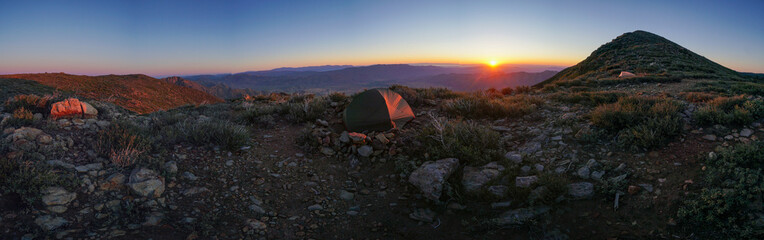 A panoramic view of a rocky mountain with a small green tent on top