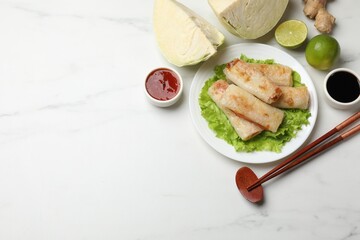 Delicious fried spring rolls, chopsticks, sauces and ingredients on white marble table, flat lay. Space for text