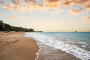 Deserted wide sandy beach with turquoise blue sea. Tropical plants of a bay at sunset in the...