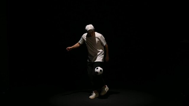 A young man throws a soccer ball on a black background. Football player shows tricks with the ball. Freestyler hits a black and white ball on a dark background. High quality FullHD footage