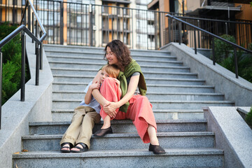 Happy family: mother and child son hugging and laugh sitting on the city stairs. motherhood...