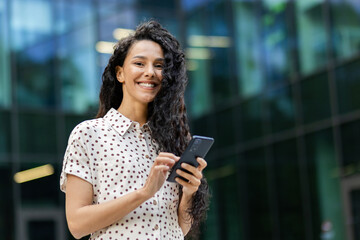 Contemporary young adult female engrossed in smartphone usage against the backdrop of a modern glass building.