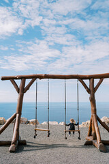 Little girl sits on a large wooden swing on a seashore and looks to the side. Back view