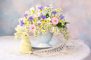 Spring bouquet with bird cherry flowers, daisies, pansies in a cup on the table, figurine of rabbit, Easter still life. - 760571742