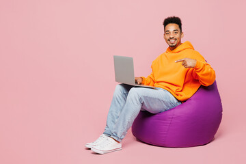 Full body young man of African American ethnicity in yellow hoody casual clothes sit in bag chair hold use work point on laptop pc computer isolated on plain pastel pink background Lifestyle concept