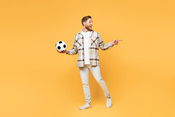 Full body young fun man fan wear brown shirt casual clothes cheer up support football sport team hold in hand soccer ball watch tv live stream point aside on area isolated on plain yellow background