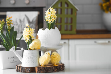 Happy Easter. Spring yellow hyacinth flowers in an egg-shaped vase, chicken chicks on a white table. In the background is a white Scandinavian-style kitchen. Easter decor in the house. - obrazy, fototapety, plakaty