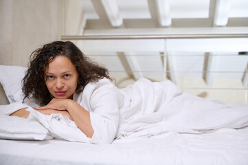 Delightful attractive pretty woman in white waffle bathrobe, relaxing on white bed sheets while waking up in the morning