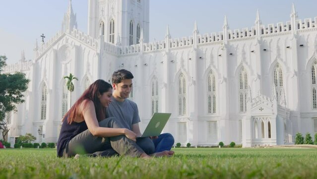A young Indian Asian cheerful attractive man and woman or romantic couple sitting outside outdoors busy using a laptop to work or study together on a university campus. education, learning concept