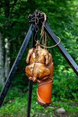 Smoked chicken and sausage on the hook and rope, hanging on the tripod over a campfire in the forest - 760621972