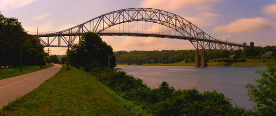 Cape Cod Canal Landscape with Sagamore Bridge at Sunrise with floating clouds and green bushes and trees along riverbank on Cape Cod, Massachusetts, USA, Long Exposure photo