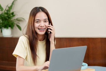 Radiant young woman with braces on call, smiling at laptop screen, room enriched with lush greenery. Beaming lady in conversation on smartphone, eyes on laptop, accompanied by vibrant potted plant.