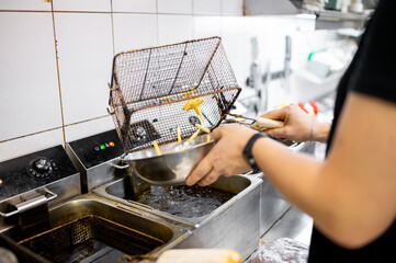person frying French fries in a commercial kitchen. They use a metal basket to lift the fries from hot oil in a deep fryer. The kitchen has white-tiled walls, and the individual wears black clothing