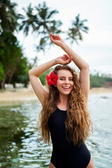 Woman enjoys serene sea swim, tropical getaway. Female in swimsuit smiles with flower in ear. Solo traveler in seaside relaxation, adventure in paradise. Oceanside retreat, a tropical beach vacation.