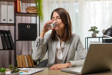 Focused young businesswoman girl sitting at workplace desk drinking water while working with laptop in home office. Lady employee freelancer looks at computer screen browse surf internet. Good habits