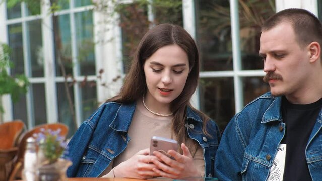 A young couple. Denim clothes. On a date in a vintage restaurant, outside. They are talking. Fooling around. They laugh funny. Taking a selfie with a smartphone.
