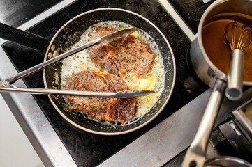 two juicy steaks sizzling in a pan with butter, being turned by tongs, next to a pot with a whisk on a stove. The steaks are well-seasoned and browning nicely, indicating they are being seared.