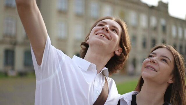 A young guy is looking for the best angle for a selfie with his girlfriend. The guy holds a mobile phone and hugs girlfriend. A couple smiles, while taking pictures against the backdrop of an old