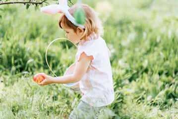 Easter egg hunt. Girl child Wearing Bunny Ears Running To Pick Up Egg In Garden. Easter tradition. Baby with basket full of colorful eggs. wide angle view - 760732925